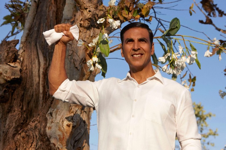 An Indian man stands in front of a tree, holding a sanitary pad triumphantly.