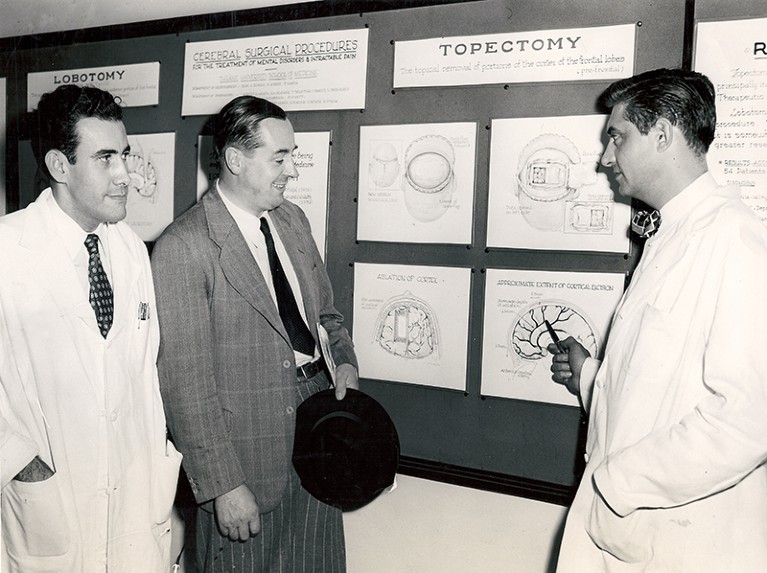 Three white men, two in lab coats and one in a suit, stand in front of a display of brain diagrams.