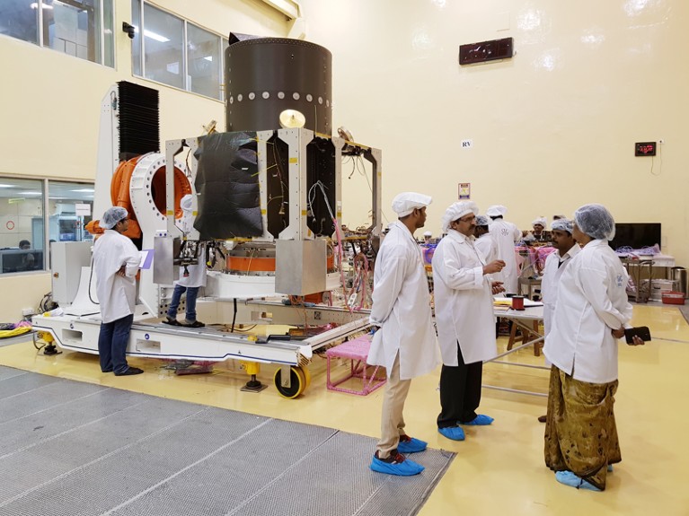 Four space engineers talk at right while other engineers work on the Chandrayaan-2 spacecraft in the background in a clean room.
