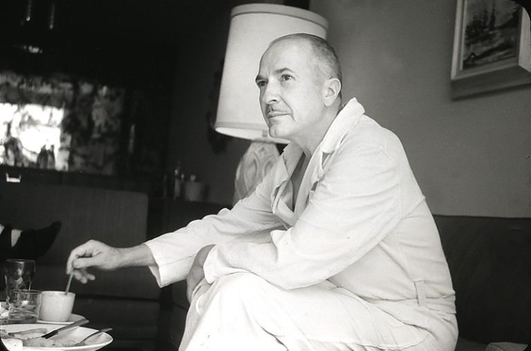 Robert Heinlein looks past the camera in his home. He is in a white dressing gown and is stirring a hot beverage.