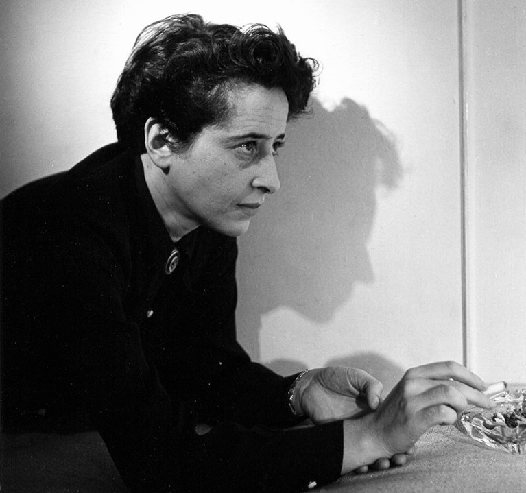 A black and white photo of Hannah Arendt in profile, leaning over a table and smoking a cigarette.
