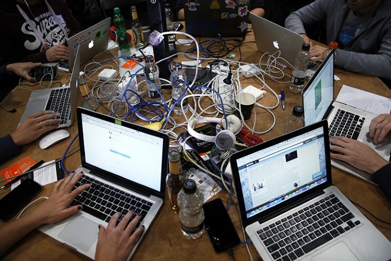Hands work at a circle of laptops on a table, surrounding a jumble of wires and equipment.