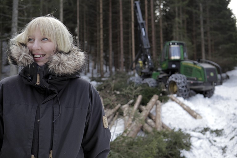 A woman in a winter coat stands in snow in front of a forest, where trees are being cut down.
