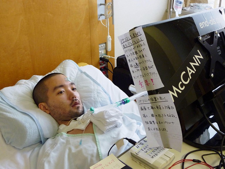 Masahiro Fujita in a hospital bed, with a breathing tube. In front of him is a monitor plastered with Japanese signs.