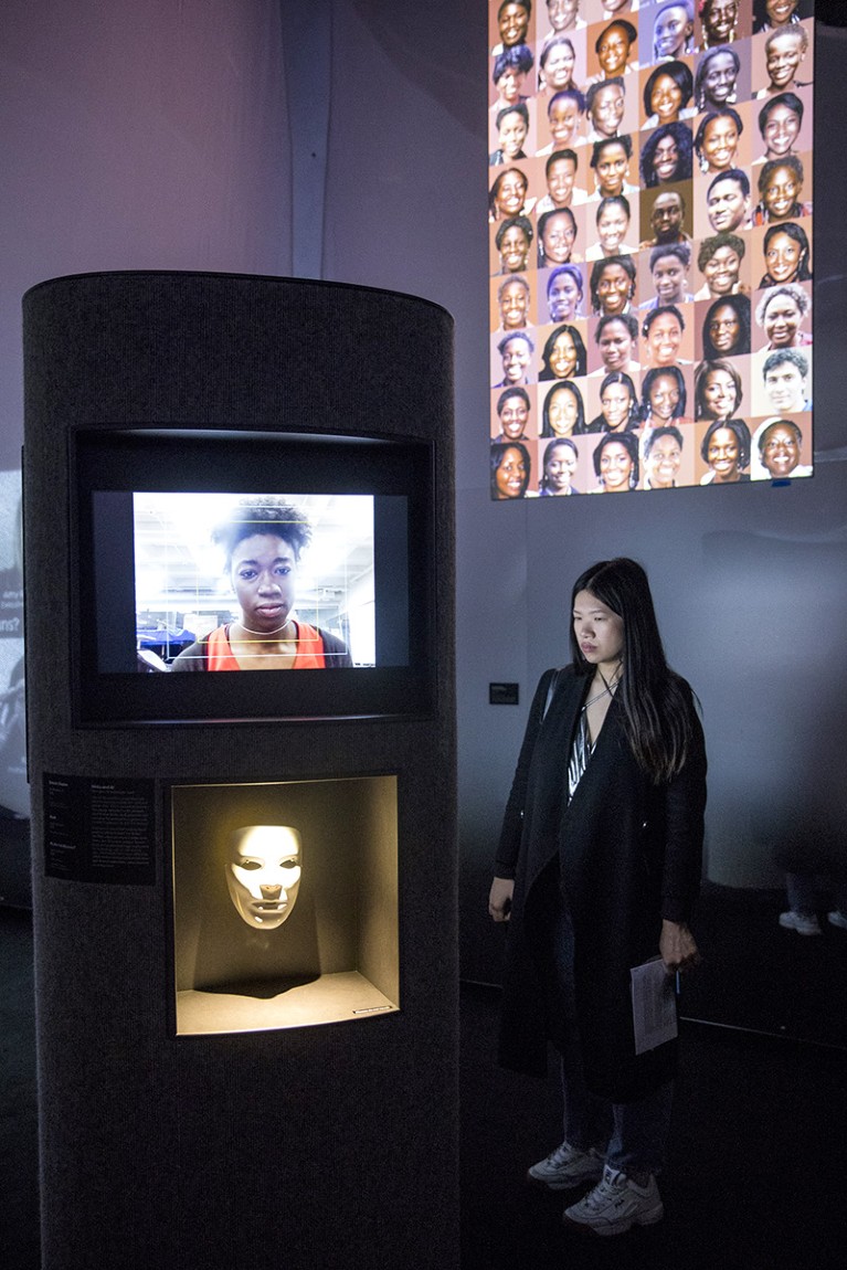 A woman looks at a TV screen showing a black woman, above a white mask, next to pictures of women of colour.