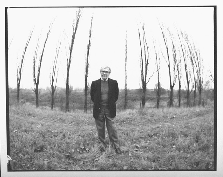 A black and white photo of James Lovelock standing in front of a row of bare trees.