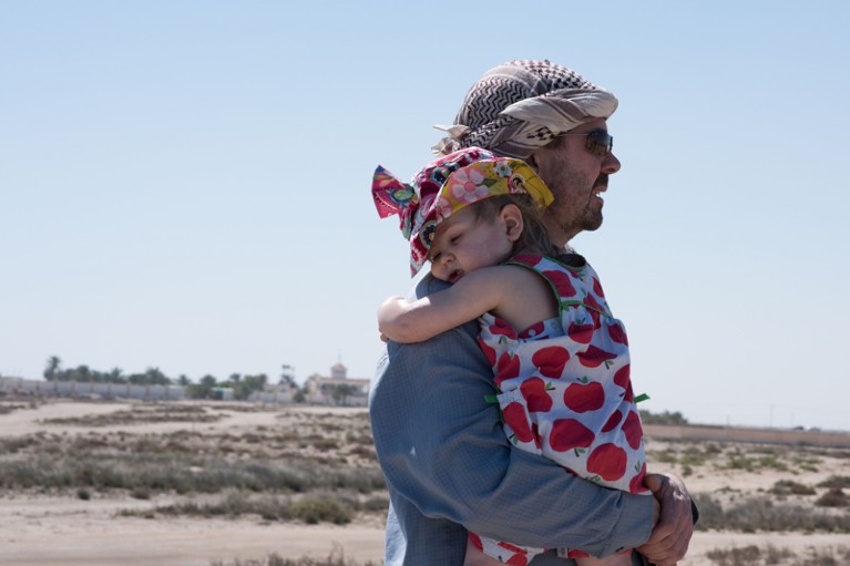 A father carries his young daughter across a desert landscape