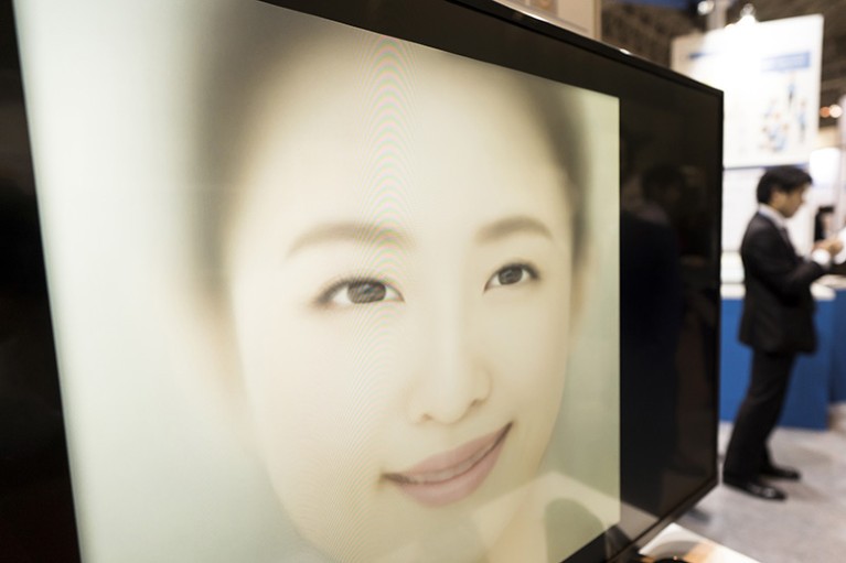 A large face on a screen, seen at an angle, with people in the background.