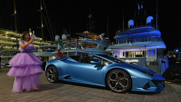A woman in a lilac ball gown standing next to a blue Lamborghini at a marina, in front of several yachts, at night.