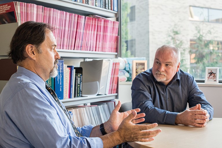 Scott Powers sits at a desk in front of a bookshelf listening to a colleague