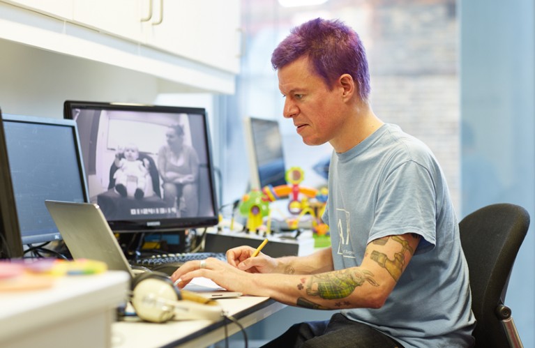 Caspar Addyman works at a desk with children's toys, headphones and monitor showing a woman and baby on the screen