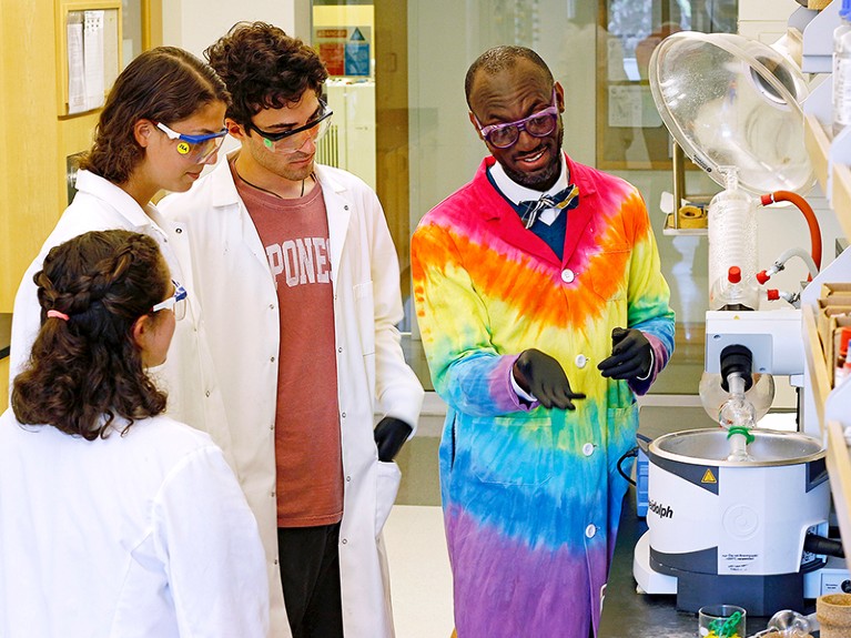 Andre Isaacs, wearing a rainbow-coloured lab coat, talks about equipment as three students look on.