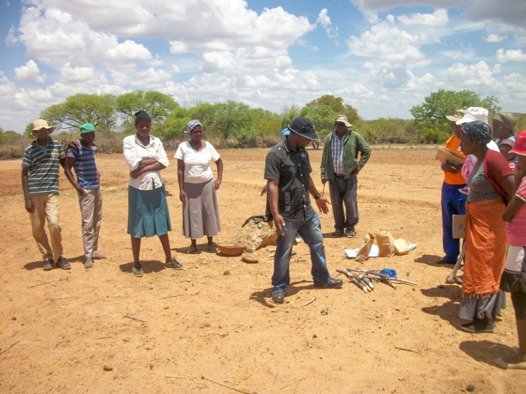 Several people standing around a man giving a demonstration in an area of parched earth, with trees in the distance.