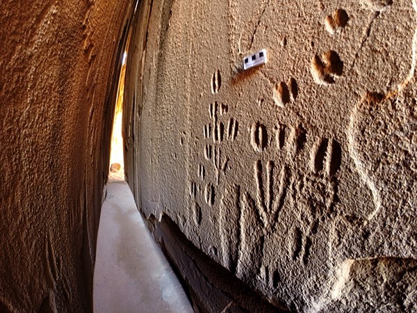 A narrow gap between two light-brown sandstone walls. The right-hand wall is covered in carvings of hoof-, foot- and paw-prints.