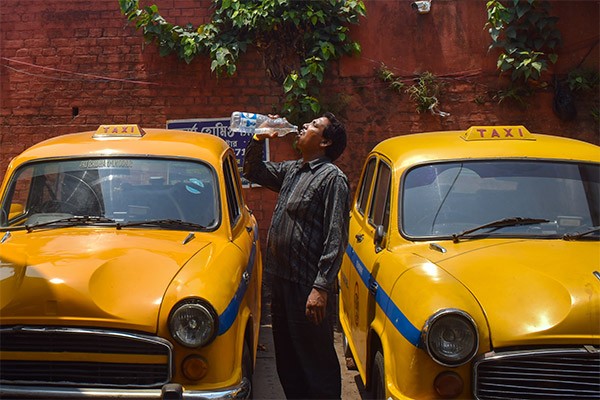 A taxi driver stood between two yellow taxis drinking water from a plastic water bottle in Kolkata, India