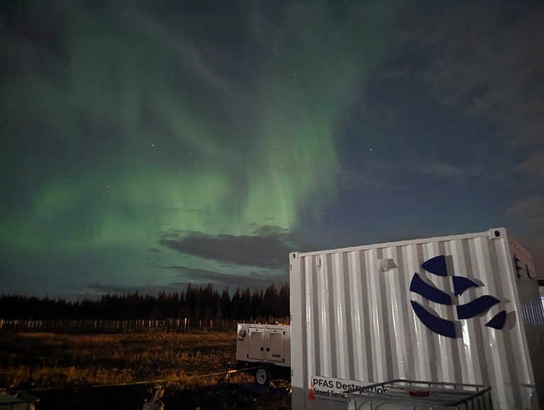 White metal shipping container in the foreground, with green Northern Lights visible in the sky above.