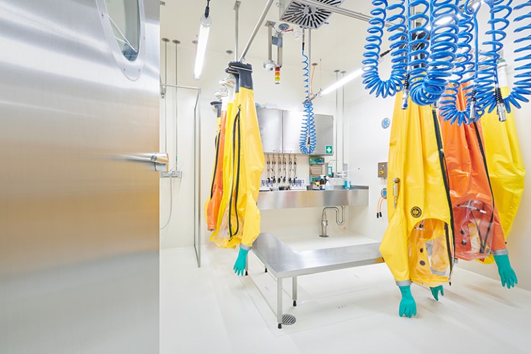 At left a door opens into a room with several yellow and orange biosafety suits handing upside from ceiling