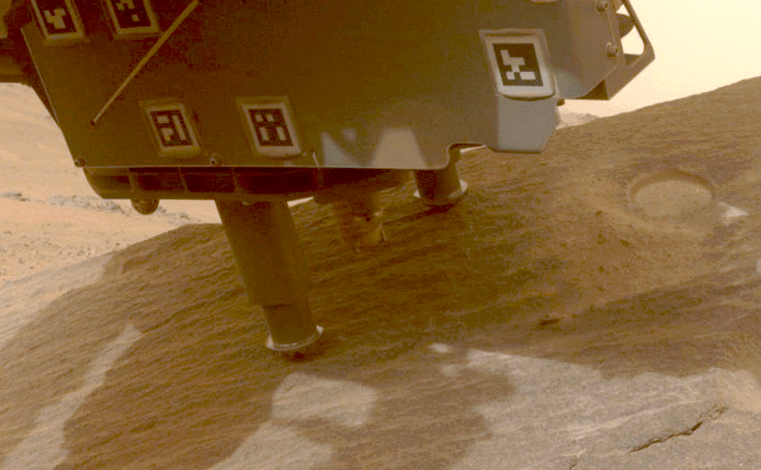 Animation of a coring bit descending from the base of a rover to drill into a sandy surface.