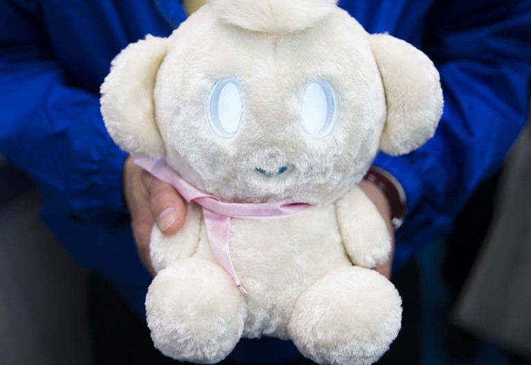 hands holding white robot with blue eyes and pink ribbon tied around neck.