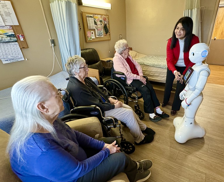 To the right person stands next to robot, facing three elderly people (one sits in a chair, the other two sit in wheelchairs)