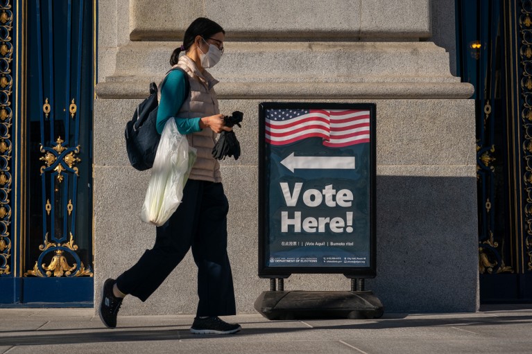 A man with a bag and a mask walks past a sign that reads “Vote Here.”