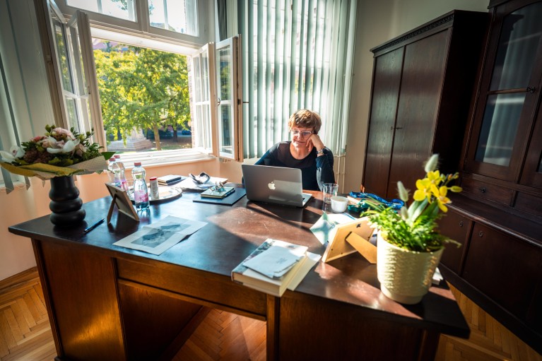 Nobel prize winner Katalin Kariko sits at a large wooden desk in an office, working on a laptop.