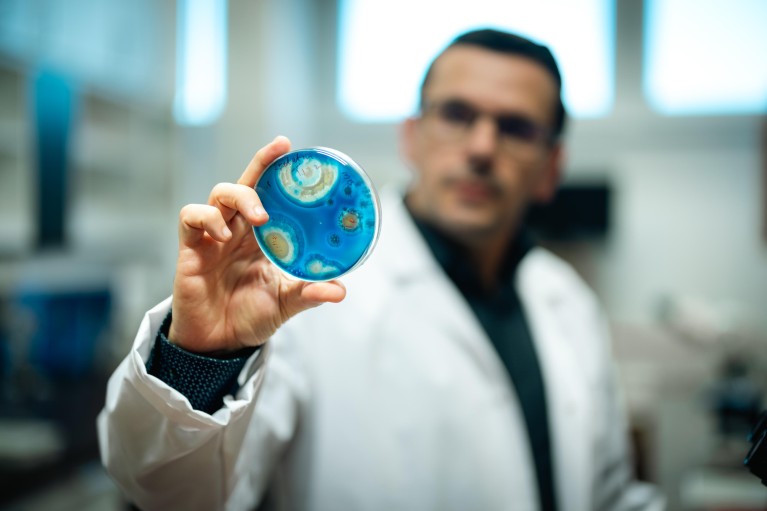 Scientist Attila Gacser, wearing a lab coat and holding a circular petri dish with a blue and yellow cell culture to the camera