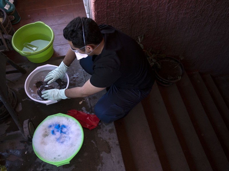 A man in mask and gloves washes his clothes in basins on the steps outside his home.