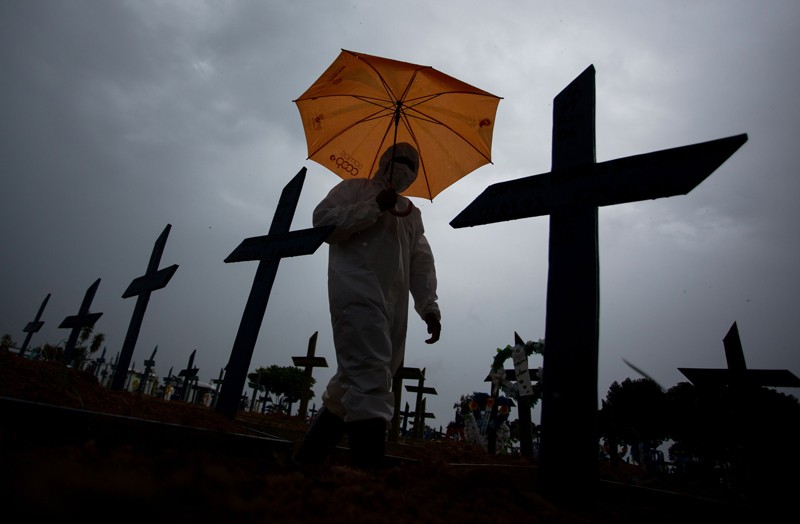 A worker wearing a protective suit and carrying an umbrella walks past the graves of COVID-19 victims, Brazil