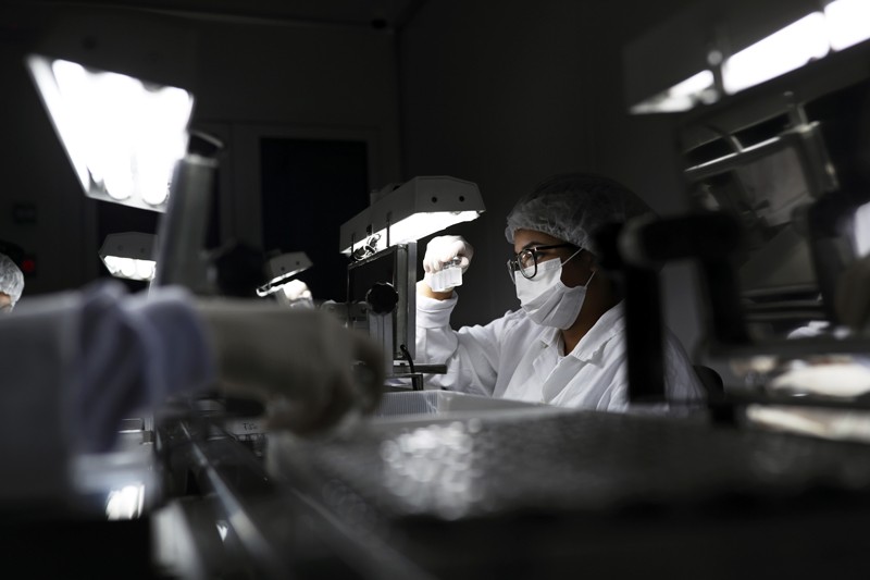 An employee wearing a protective mask and hair net inspects vials containing a COVID-19 vaccine under a light