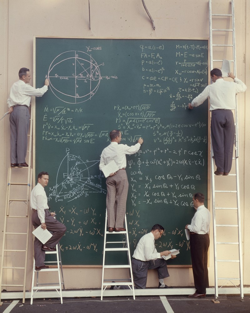 Six scientists on ladders write equations on a large chalk board.