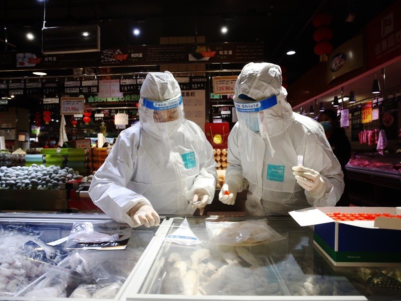 Two people in protective gear and visors swab food in a freezer at a market.
