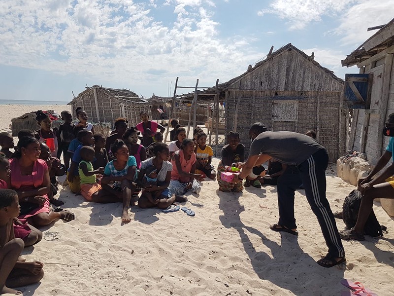 A man hands supplies to people seated on sandy ground in front of wooden houses.