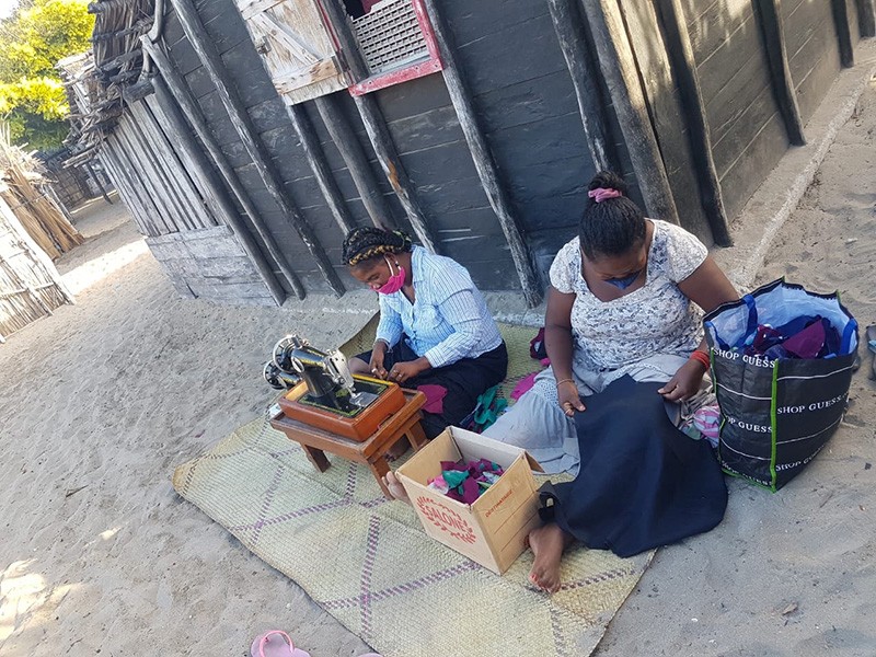Womena sit on a mat on sandy ground. One is sewing with a sewing machine, the other is handling cloth.