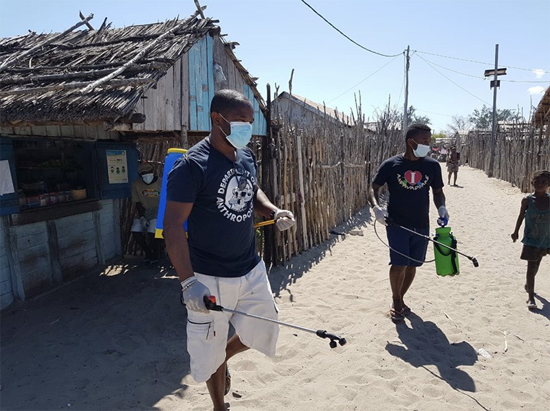 Two men spray disinfectant in a town with wooden buildings and sandy ground.