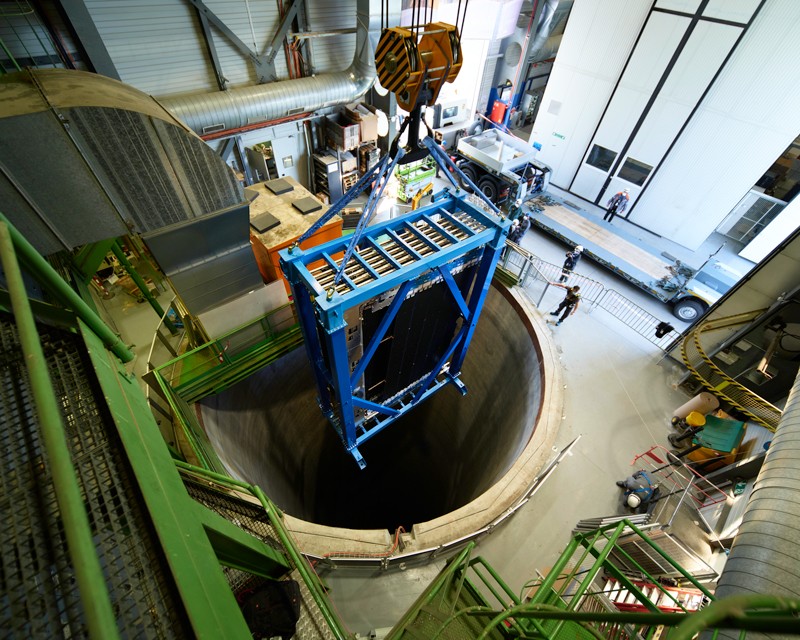 A large detector module that makes up the LHCb is lowered by a crane into a shaft in the floor of a warehouse