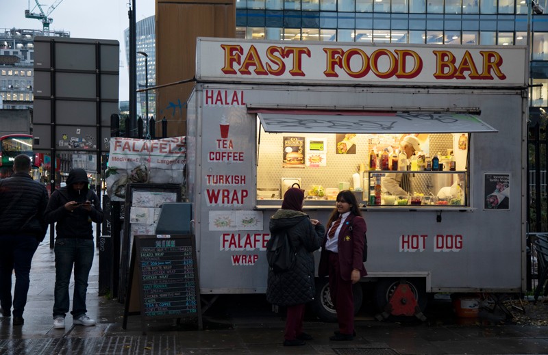 Schoolchildren stand next to a fast food van as a man nearby smokes a cigarette in East London