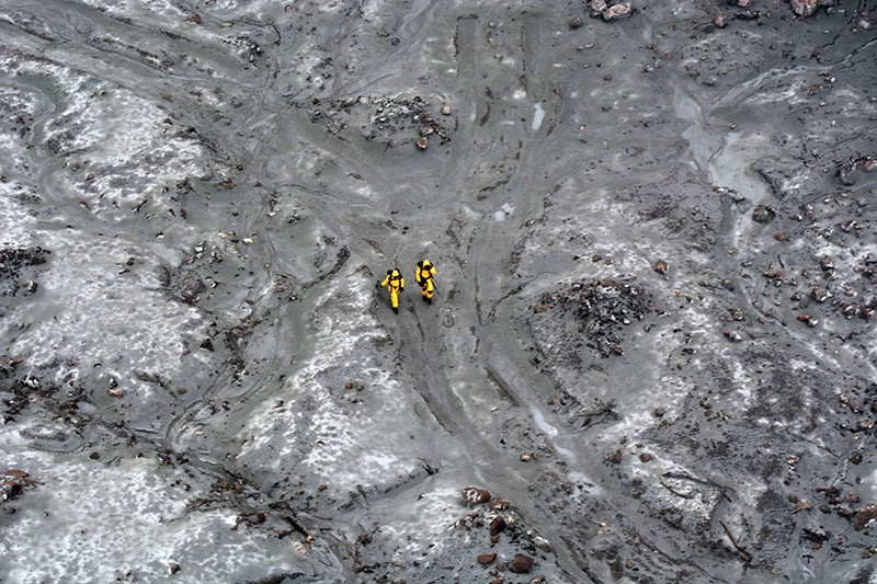 Two figures in yellow hazmat suits walk across the devastated landscape of Whakaari/White Island.
