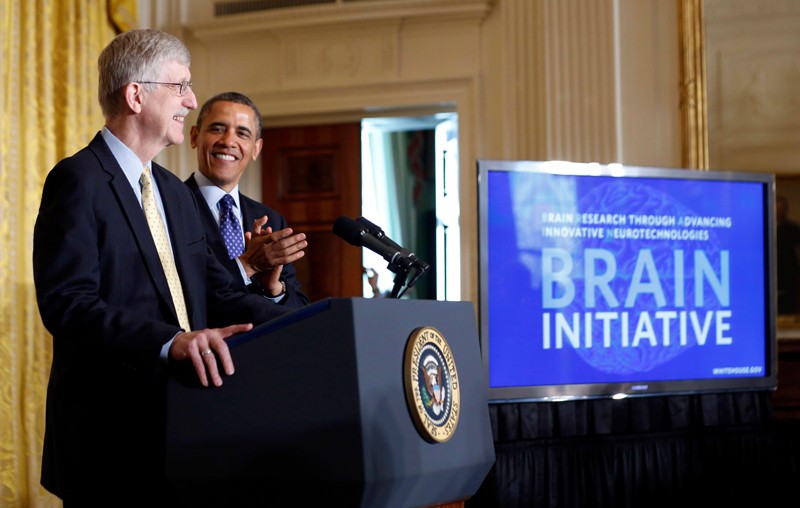 Francis Collins and Barak Obama stand at a podium at a press conference beside a monitor displaying the words 'Brain Initiative'