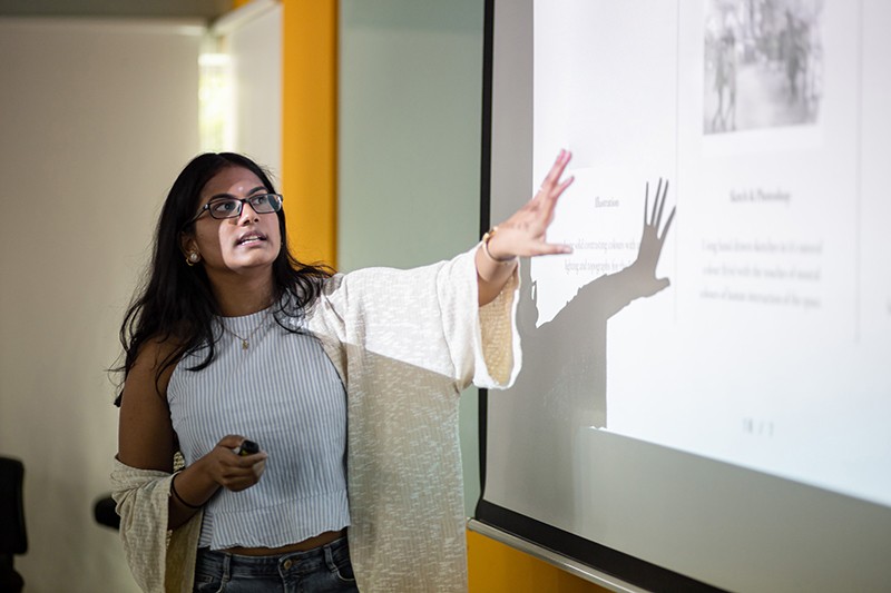 A researcher explains a slide while holding her arm out towards a presentation projected on a whiteboard.