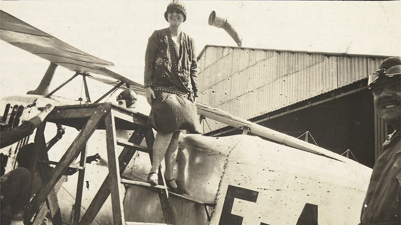 Bertha Lutz stands on a ladder next to an airplane with a pilot in the front while people look on.