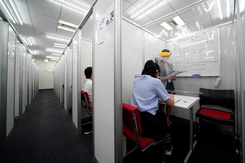 An instructor wearing PPE teaches a student through a plastic barrier in a cubicle at an education centre in Japan