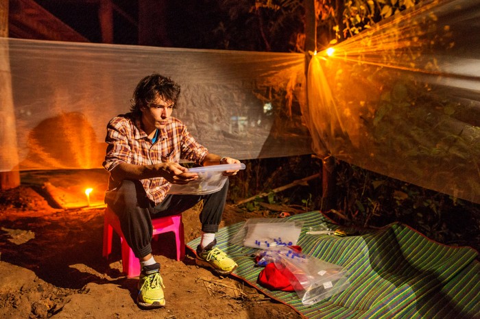 Victor Chaumeau sits outdoors at night with bags of plastic vials, mosquito nets suspended around him and candles burning