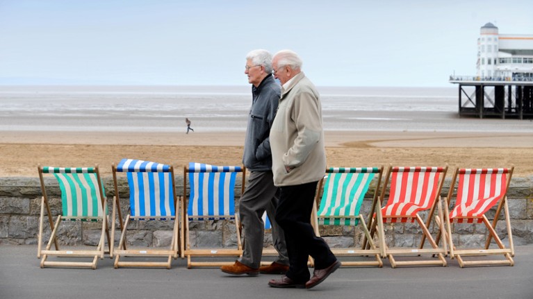 Two older men walk past deckchairs lined up next to a wall by a beach.