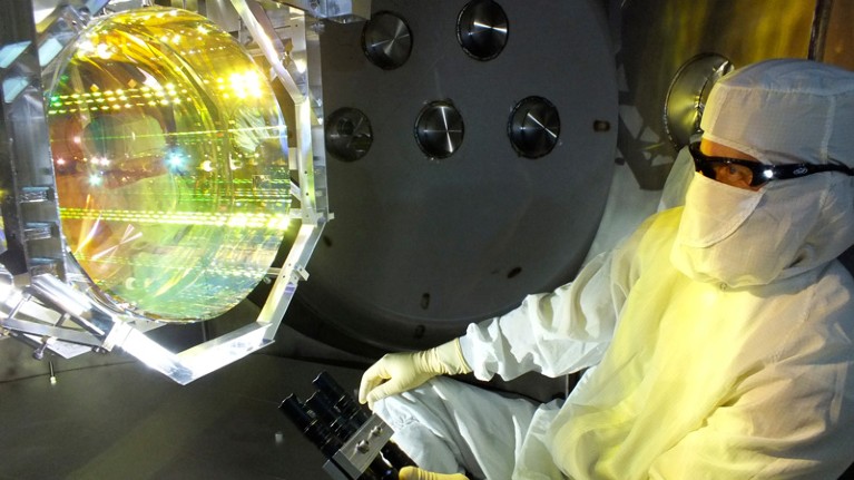 A person in a clean-room suit and goggles looking at an optical device a little bigger than their head.