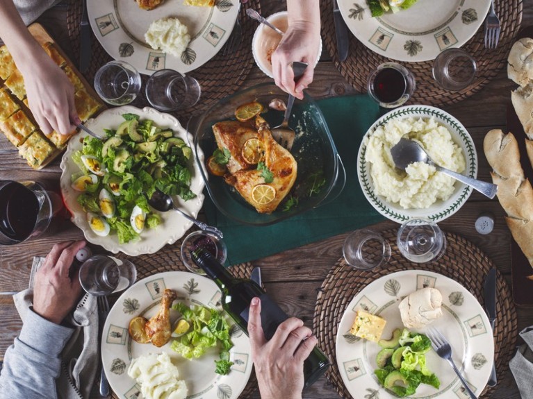 Overhead shot of lots of full plates on a table, with hands reaching to take food or pour wine.