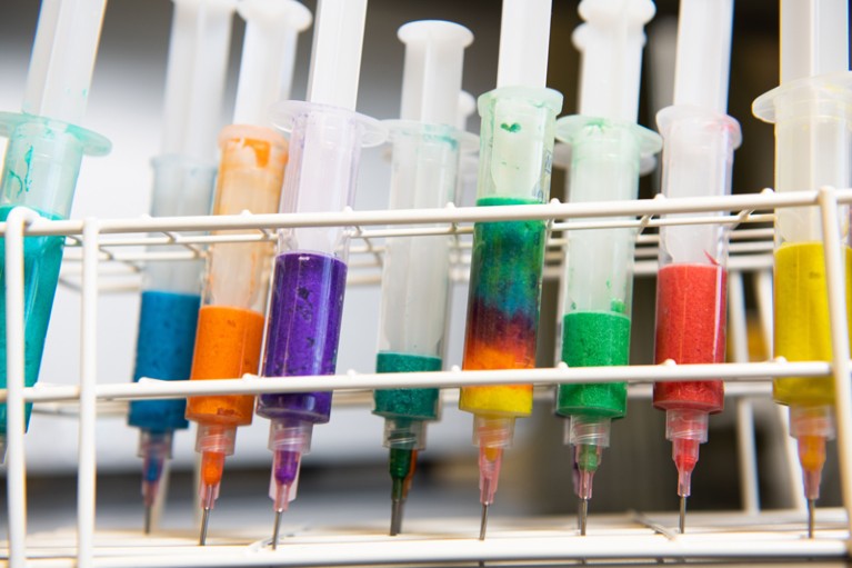 Syringes filled with various brightly coloured liquids stand in a test-tube rack.