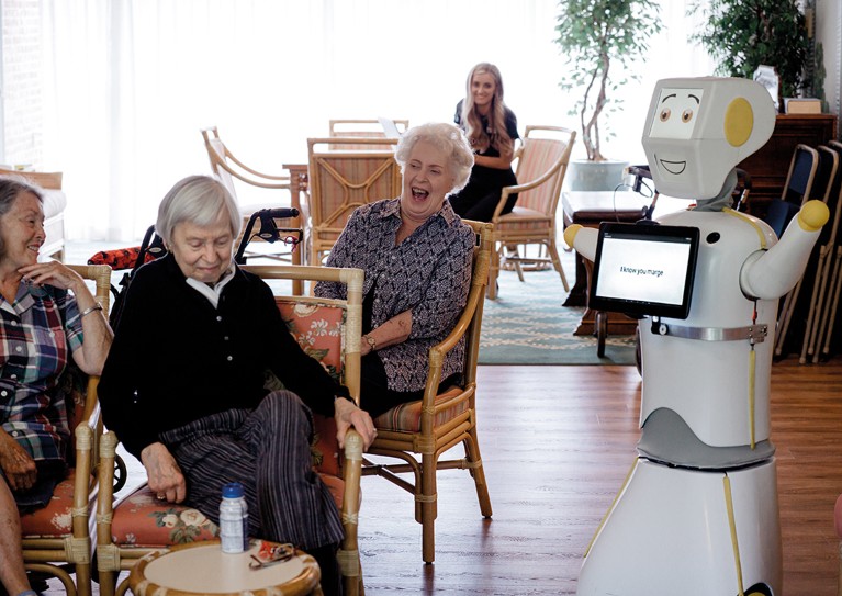 A robot stands in front of elderly people sitting in chairs and laughing