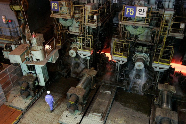 A high-angle view of a worker wearing a helmet standing beside a hot strip mill inside a factory of steel