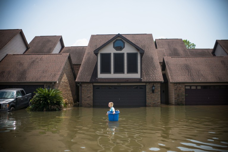 A woman wades through flood water past a house while carrying a large blue bucket.